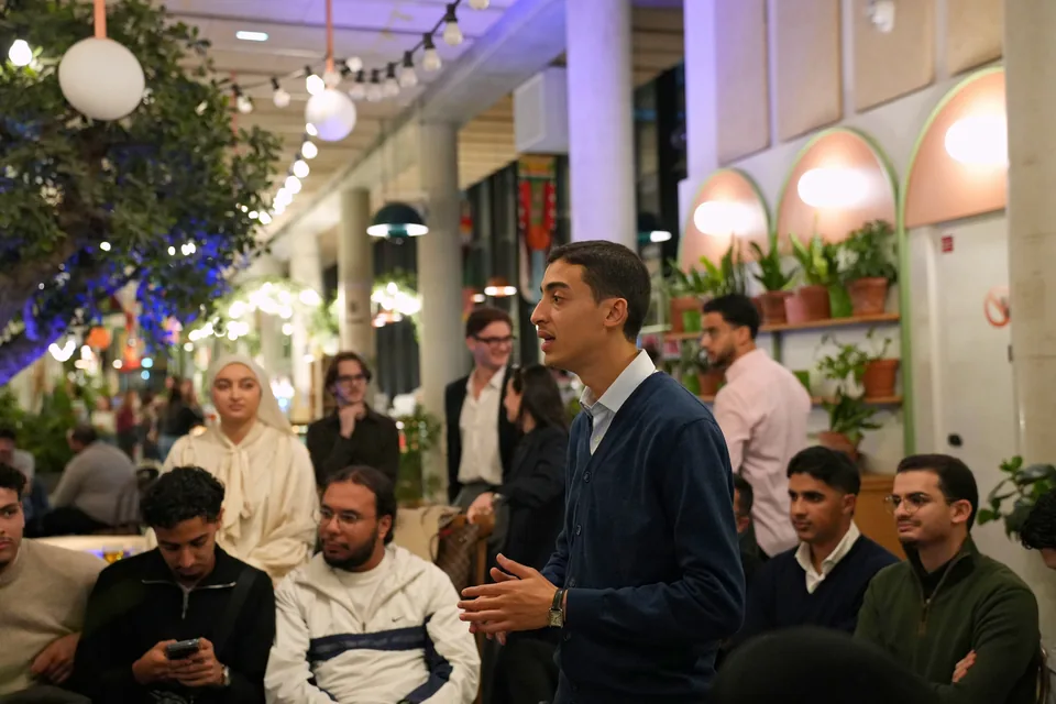 A young man in a navy cardigan speaks to a seated audience beneath strings of lights and illuminated arches.