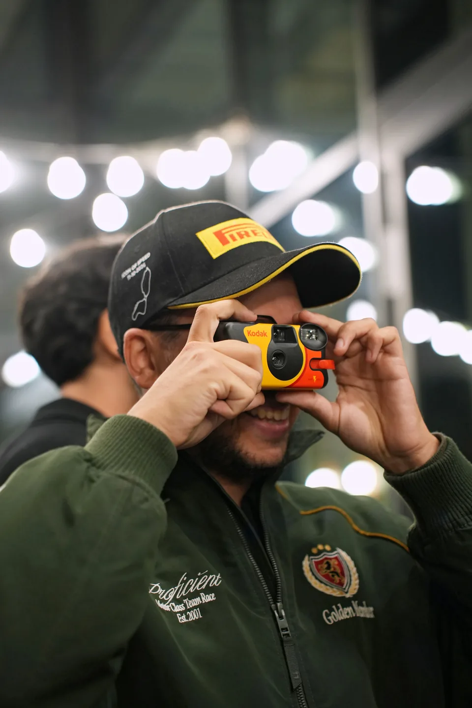 A man in a cap and green jacket aims a yellow disposable camera at the lens, soft lights behind him.